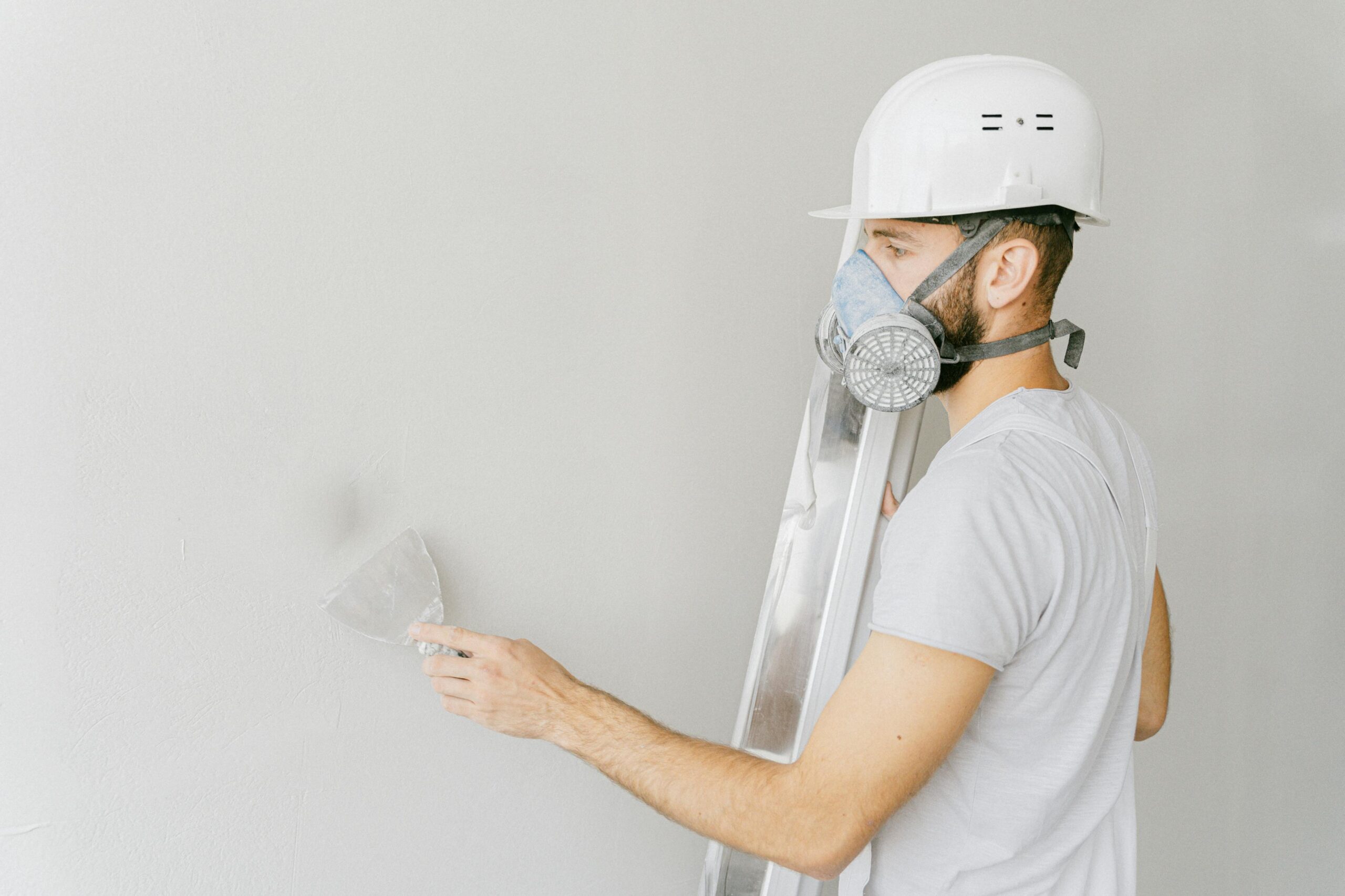A construction worker wearing a gas mask and hard hat applies plaster to a wall indoors.